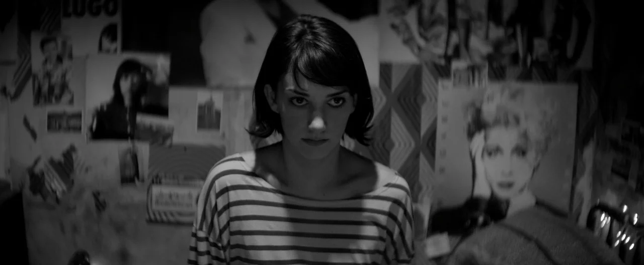 A Girl Walks Home Alone at Night (2014) movie still: medium — A dimly lit room features walls covered with various paper posters and photographs. A…