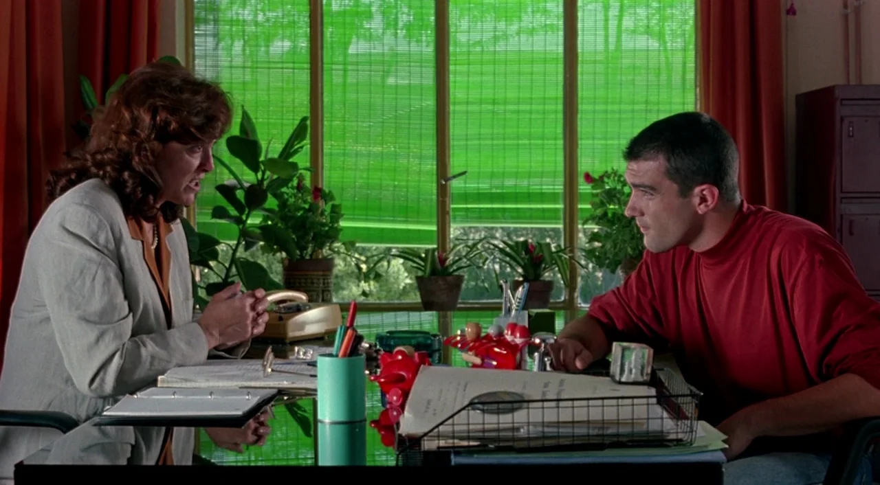 Tie Me Up! Tie Me Down! (1989) movie still: medium — An office with a glass desk, green bamboo blinds, and several potted plants. A woman in a grey…
