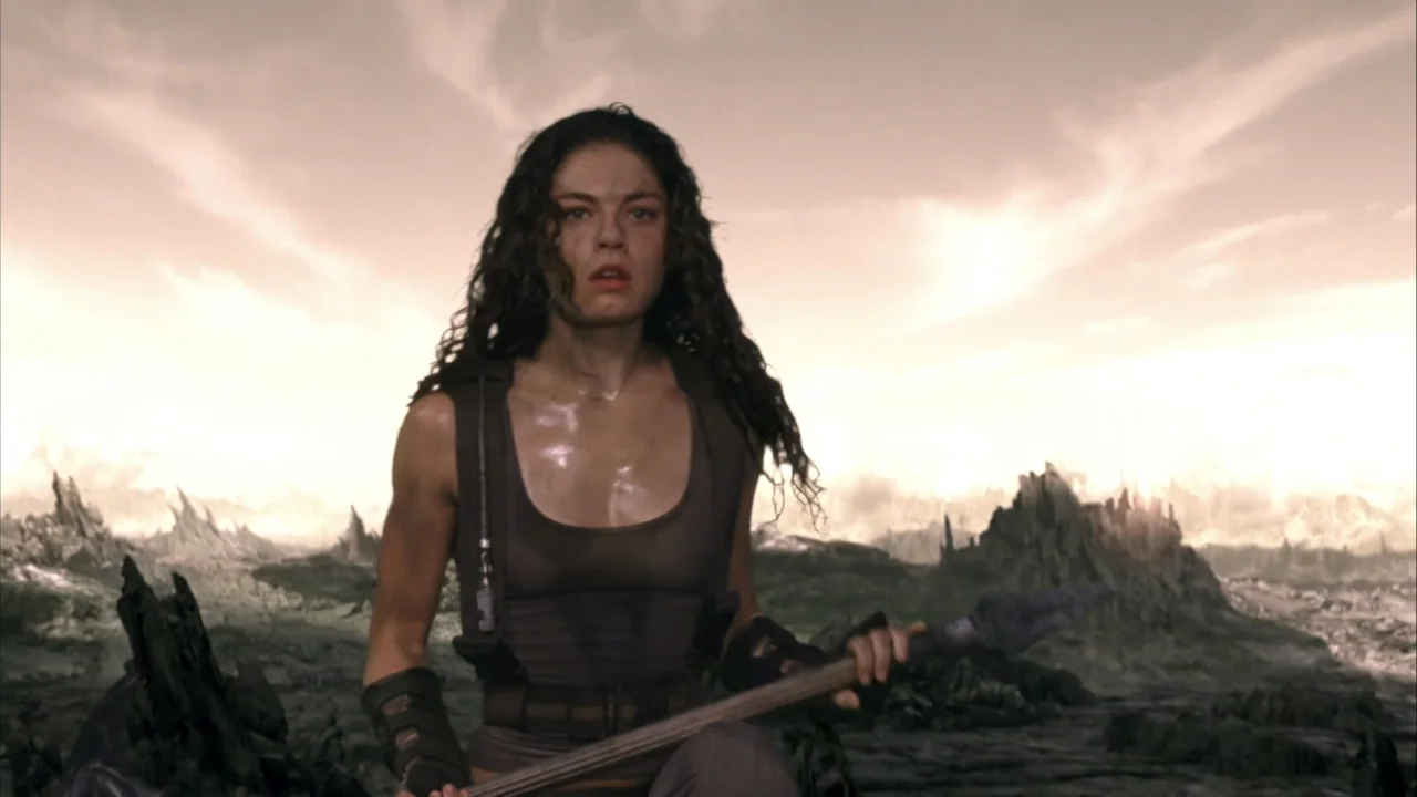 The Chronicles of Riddick (2004) movie still: medium — A desolate and rocky landscape stretches out under a pale, overcast sky. A woman with dark…