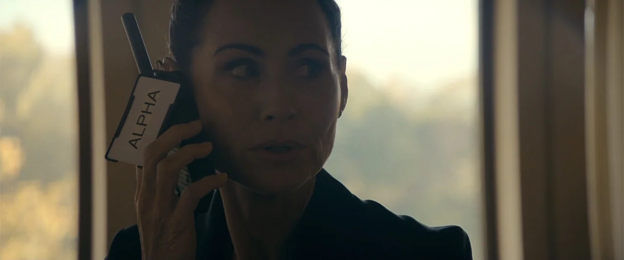 The Beekeeper (2024) movie still: closeup — An interior office space with a bright window visible in the blurred background. A woman in a dark suit…