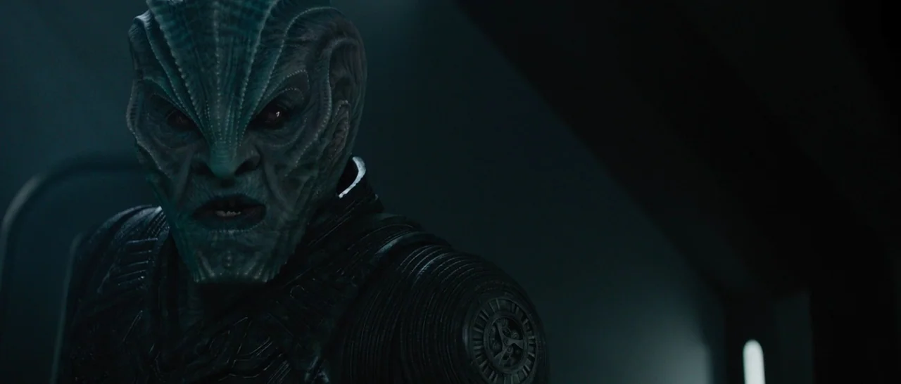 Star Trek Beyond (2016) movie still: closeup — A dimly lit interior space with dark, textured wall panels and shadow-filled corners. An alien with…