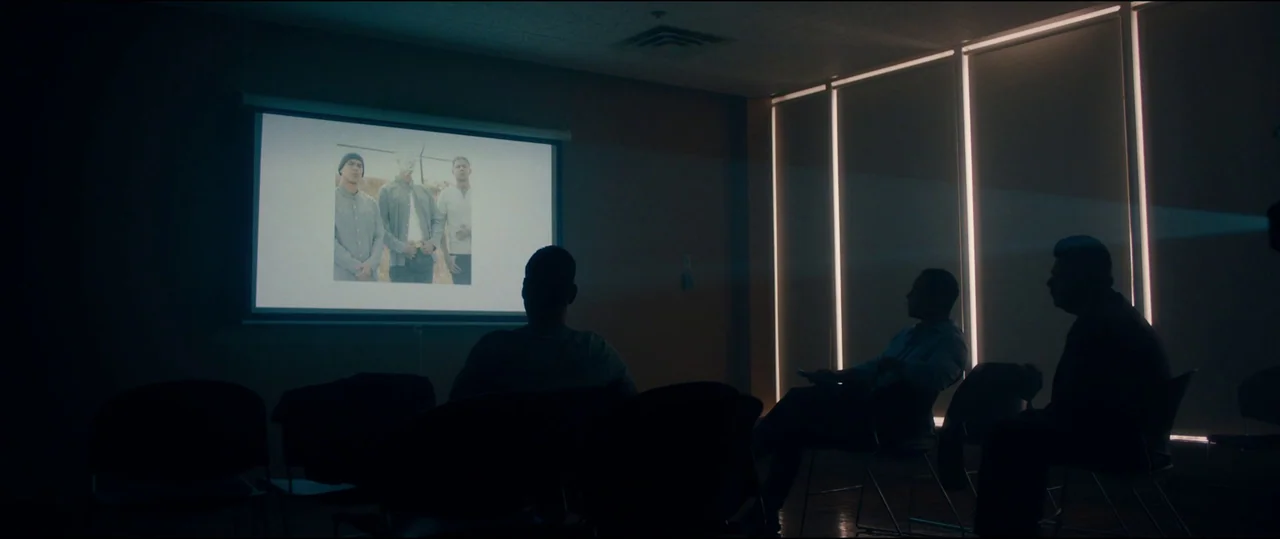 El Chicano (2018) movie still: wide — A dark conference room contains several chairs arranged in front of a projection screen. Three men sit facing…