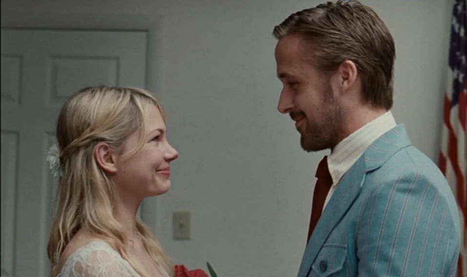 Blue Valentine (2010) movie still: medium — A spartan room with off-white walls features a closed door and an American flag. A young bride and groom…
