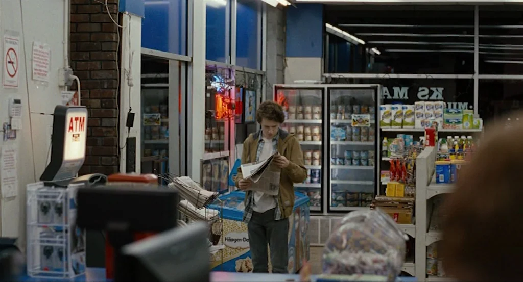 Louder Than Bombs (2015) movie still: medium — A well-lit convenience store interior features rows of products and glass-front freezer units. A…