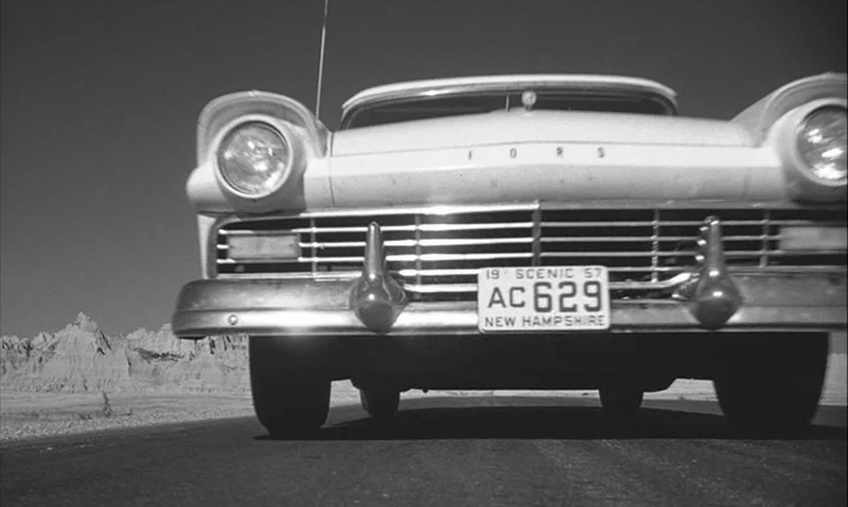 Lolita (1962) movie still: wide — A 1950s Ford sedan drives down a paved road in a rocky, arid landscape. The camera is positioned low to the ground…