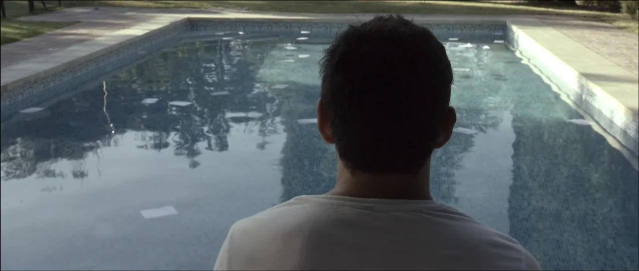 Otro verano (2013) movie still: medium — A man stands before a rectangular swimming pool with clear water and stone coping. Numerous white papers…