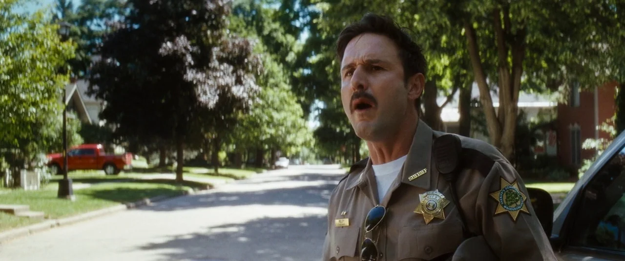 Scream 4 (2011) movie still: medium — A suburban residential street is lined with mature trees and houses under daylight. A sheriff stands in the…