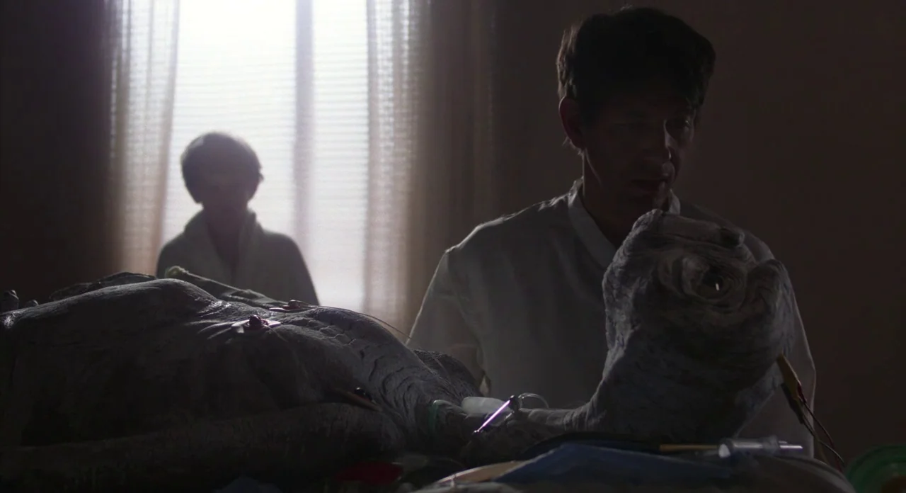 E.T. The Extra-Terrestrial (1982) movie still: medium — A dimly lit room functions as a makeshift medical area with an alien creature. A man in a…