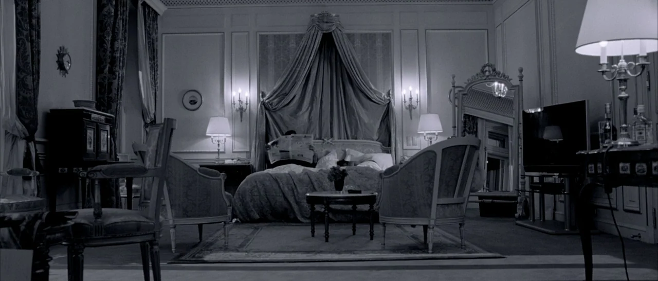 Angel-A (2005) movie still: wide — A luxuriously appointed hotel suite features a central canopy bed and ornate furniture pieces. An individual sits…