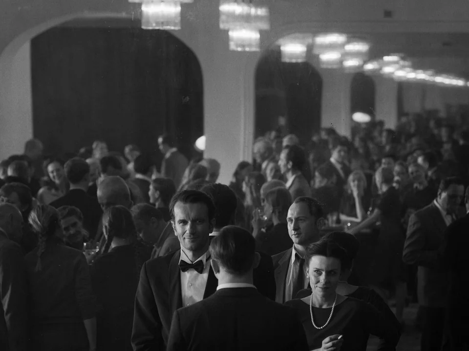 Cold War (2018) movie still: medium — A crowded ballroom with arched structural columns and multiple hanging crystal chandeliers overhead. Numerous…