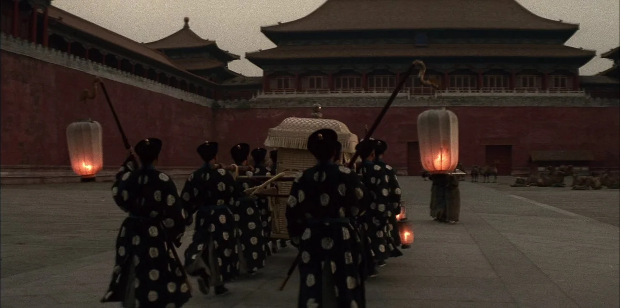 The Last Emperor (1987) movie still: wide — A group of men in traditional tunics carry a covered sedan chair across a courtyard. Behind them stands…