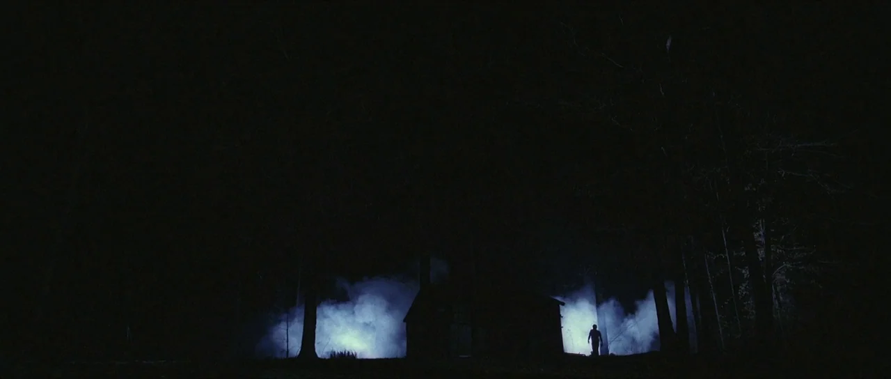 The Mothman Prophecies (2002) movie still: wide — A dark wooden structure stands in a dense, moonlit forest at night. A lone silhouette figure walks…