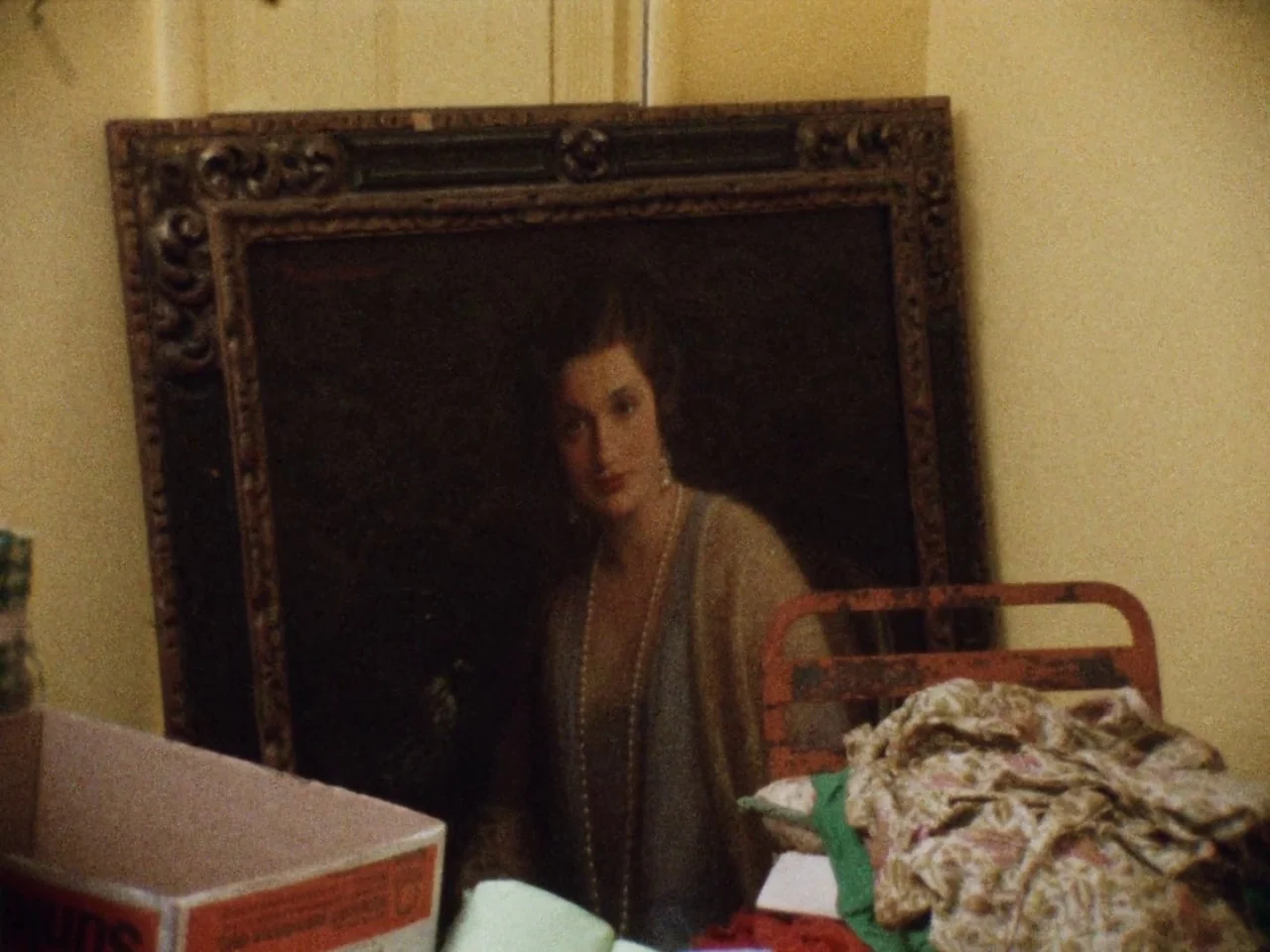 Grey Gardens (1975) movie still: medium — A framed portrait of a 1920s woman leans against a yellow residential wall. The artwork is positioned next…