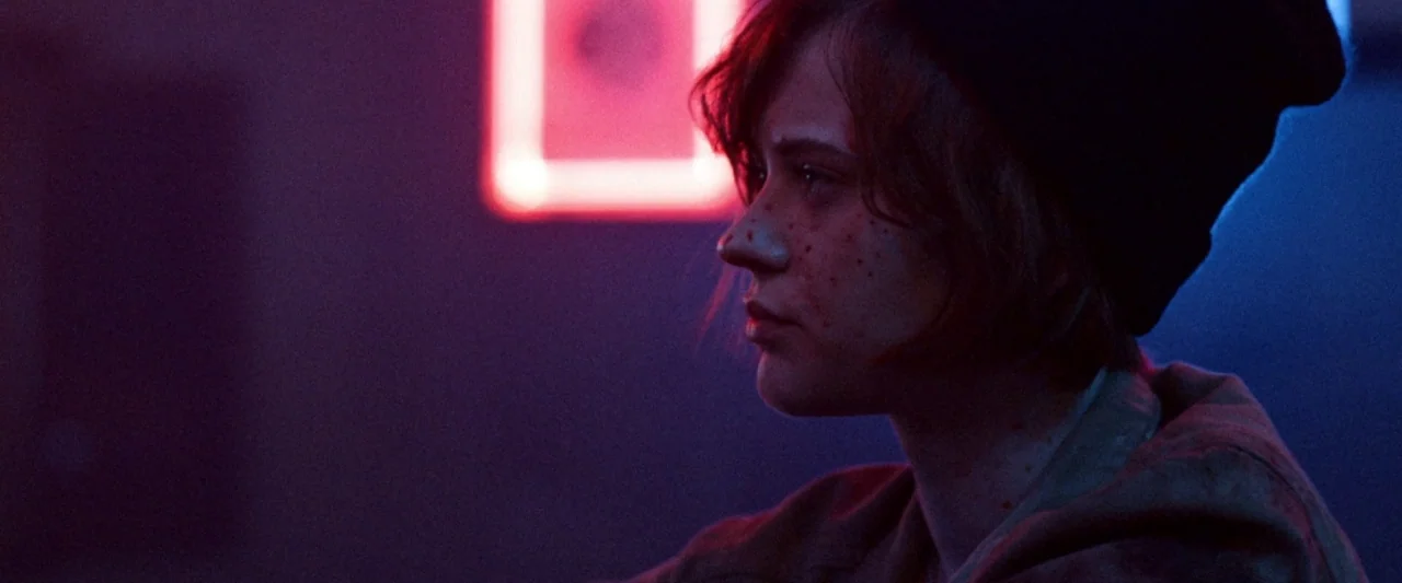 VFW movie still: closeup — A dimly lit club interior features glowing red neon signage in the background. A young woman with short hair and a beanie…
