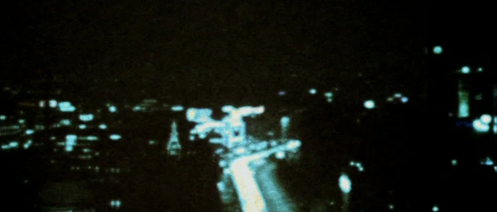 Reconstruction (2003) movie still: establishing — A high-angle night view of an out-of-focus city with blurred streaks of light. The dark scene is…