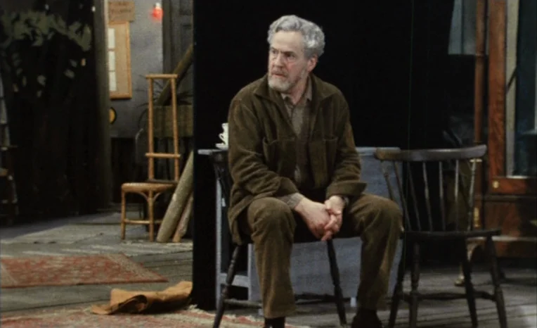 After The Rehearsal (1984) movie still: medium — A man sits on a wooden chair within a sparsely decorated studio space. Black partitions stand…
