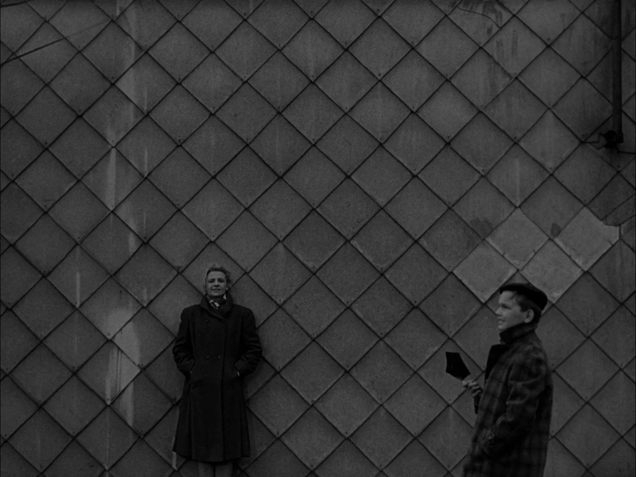 The Sun In A Net (1963) movie still: wide — A woman stands against a diamond-patterned metal tiled wall. A boy in a plaid coat observes her from the…