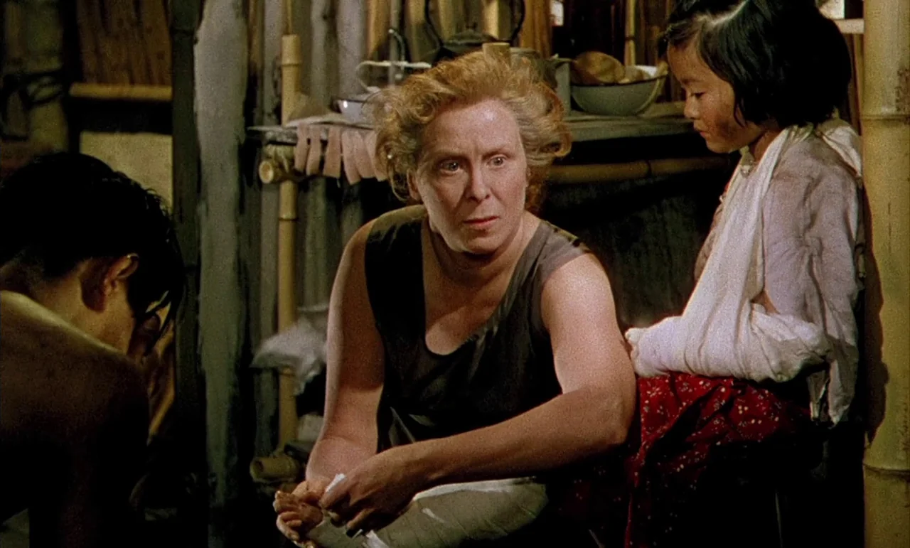 The Purple Plain (1954) movie still: medium — A makeshift structure with bamboo walls and cluttered domestic items in the background. A woman sits…