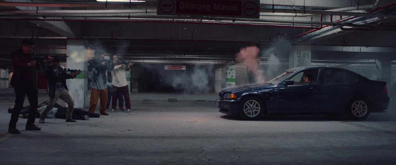 The Night Comes for Us (2018) movie still: wide — A concrete multi-level parking garage features red signage and pipes mounted on the ceiling. A…