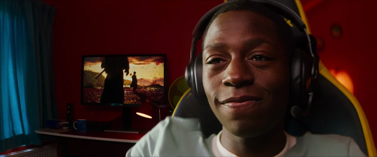 Rye Lane (2023) movie still: closeup — A dimly lit room features red walls and a computer desk with a gaming chair. A young man sits in the gaming…