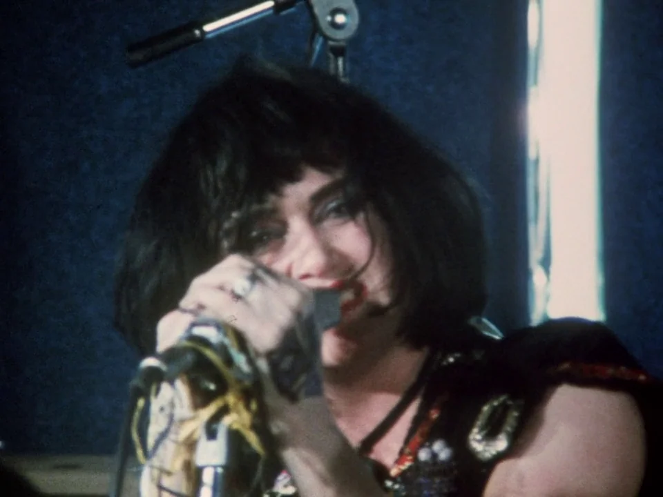 Decline of Western Civilization (1981) movie still: closeup — A dark indoor space with a sound-dampened wall and a microphone stand. A woman with…