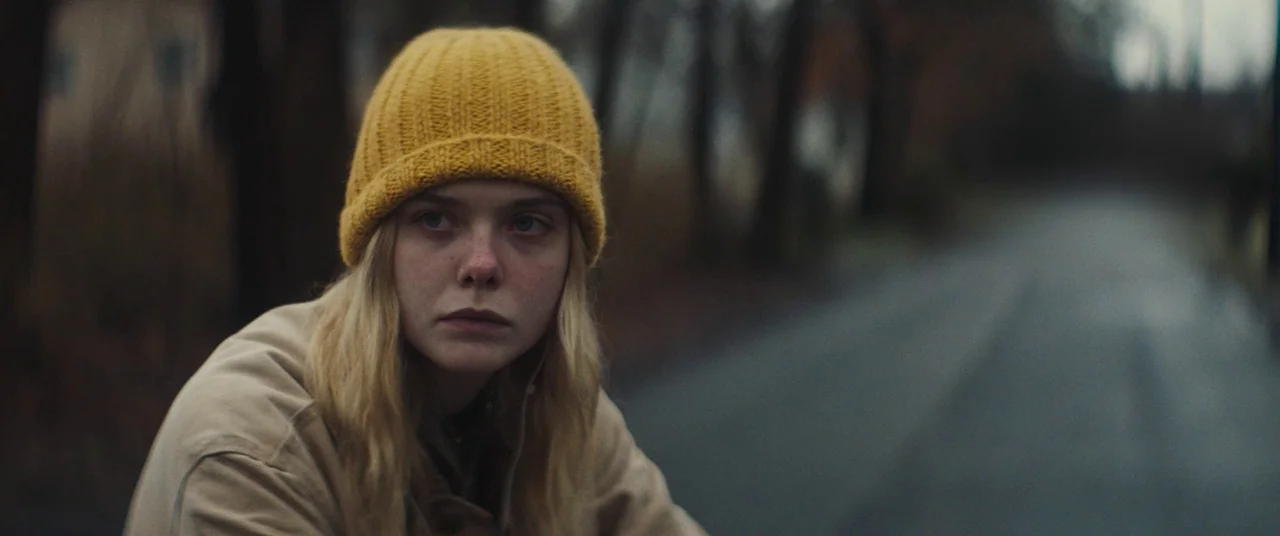 I Think We’re Alone Now (2018) movie still: medium — A rural asphalt road lined with dark, leafless trees under a gray overcast sky. A young woman…