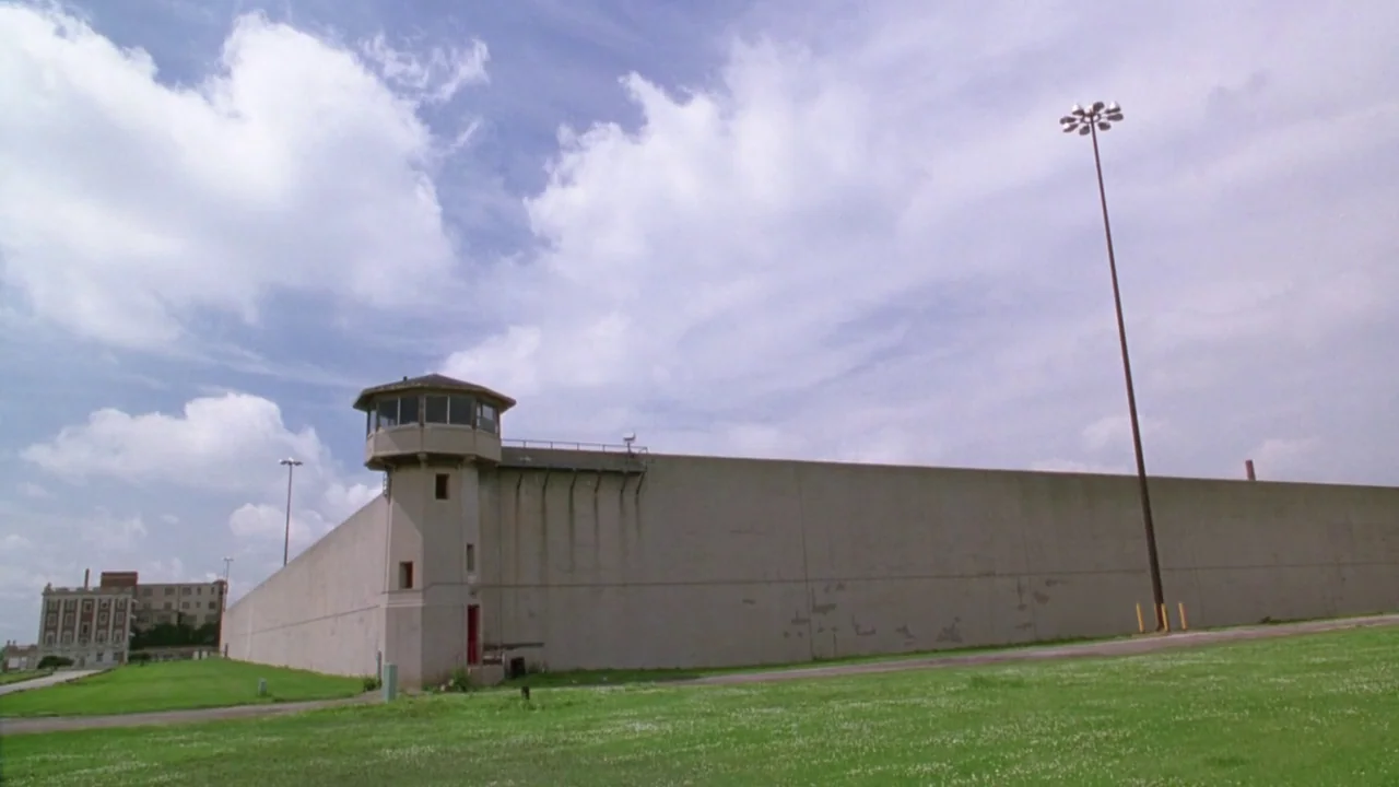 Natural Born Killers (1994) movie still: establishing — A high, smooth concrete perimeter wall stretches across the frame with a guard tower. An…