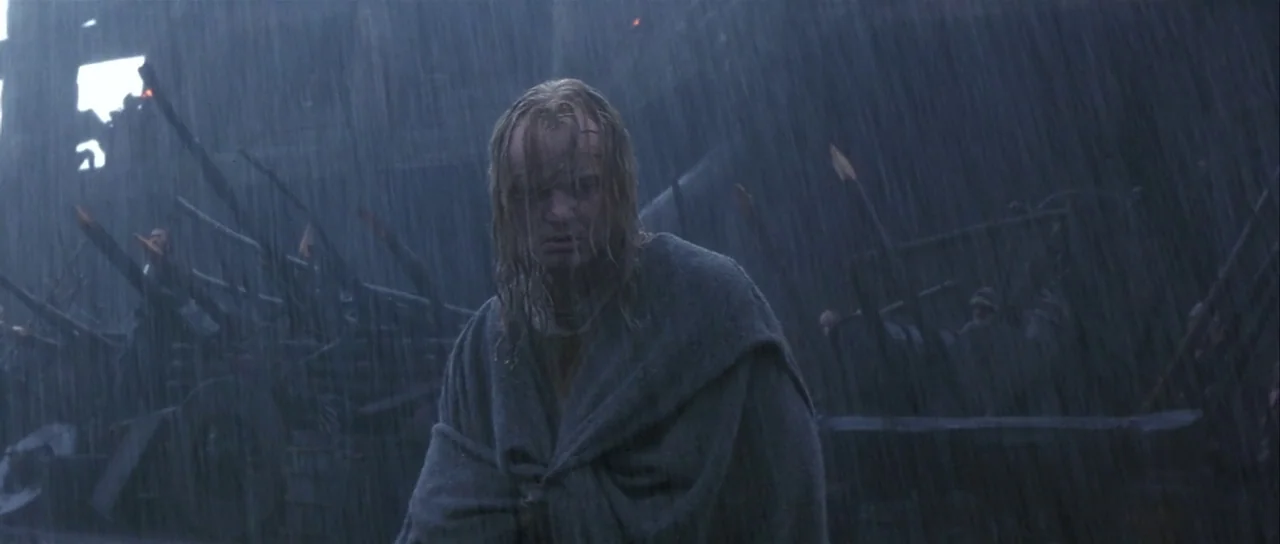 The 13th Warrior (1999) movie still: medium — A rain-drenched, dark battlefield features wooden siege weaponry and debris in the background. A man…