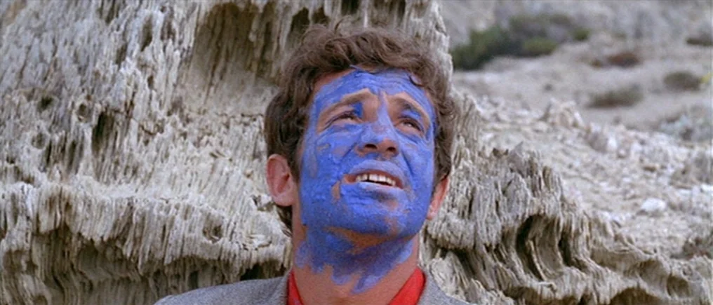 Pierrot Le Fou (Crazy Pete) (1965) movie still: closeup — A man stands in front of rough, weathered limestone rock formations. He has blue paint…