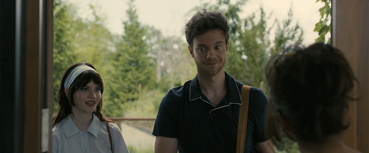Companion (2025) movie still: medium — A man and woman stand in the threshold of an open doorway. The man smiles while looking at an unseen person…