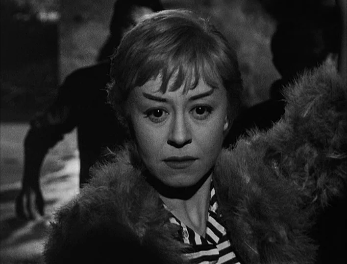 Nights of Cabiria (1957) movie still: closeup — A woman stands in a dark urban environment with out-of-focus figures in the background. She faces…