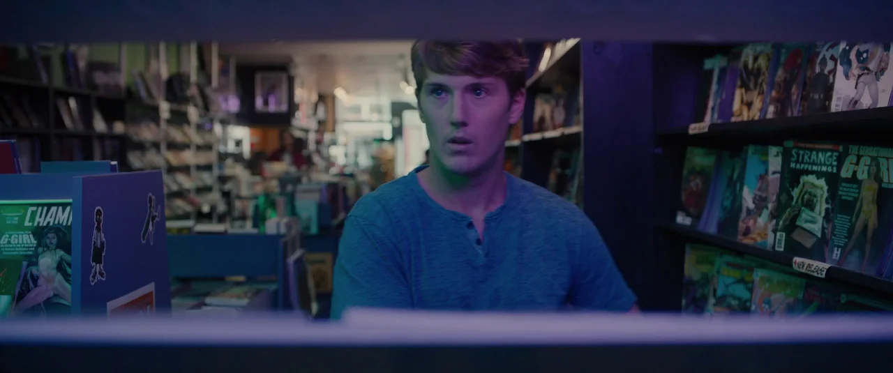 Glass (2019) movie still: medium — A comic book store interior features shelves packed with magazines under soft lighting. A young man sits in the…