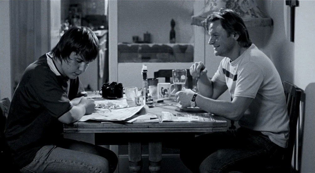 Somers Town (2008) movie still: medium — A wooden dining table features a newspaper, a camera, and various mugs. A teenager sits across from a…