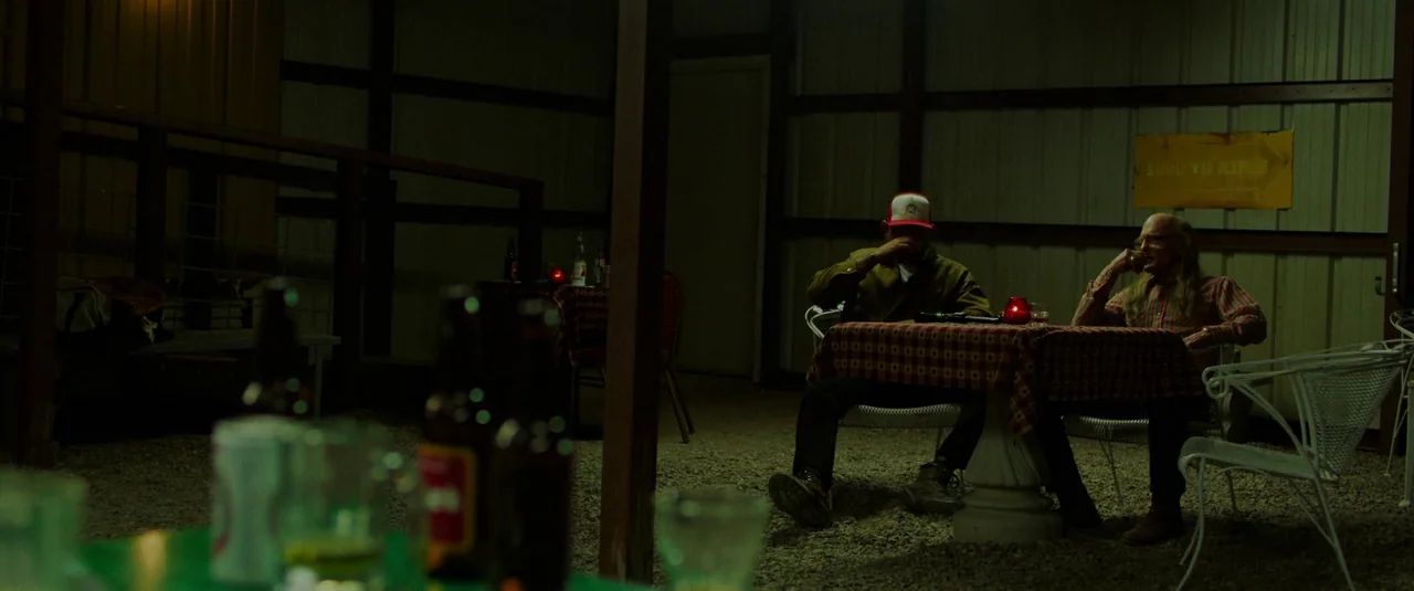 Love Lies Bleeding (2024) movie still: wide — Two men sit at a small table inside a dim corrugated metal shed structure. Gravel covers the floor…