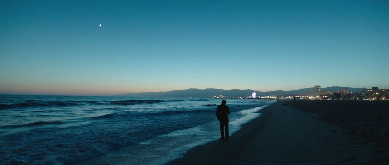 Knight of Cups (2015) movie still: wide — A dark coastline at dusk features a distant city skyline with a glowing ferris wheel. A lone figure in a…