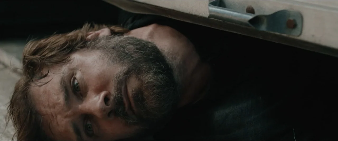 Possessions (2024) movie still: closeup — A close-up view from beneath a vehicle looking at a man lying on pavement. The metallic chassis of the car…