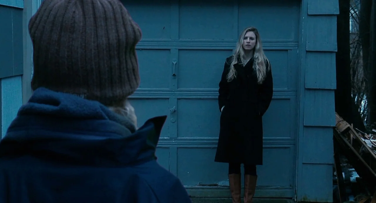 Another Earth (2011) movie still: medium — A woman stands before the blue painted panels of a closed residential garage. A person wearing a brown…