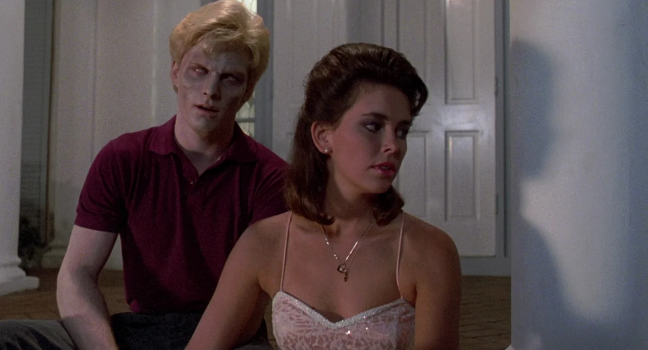Night of the Creeps (1986) movie still: medium — Two individuals sit on a porch area outside a large white house at night. A pale-skinned man with…