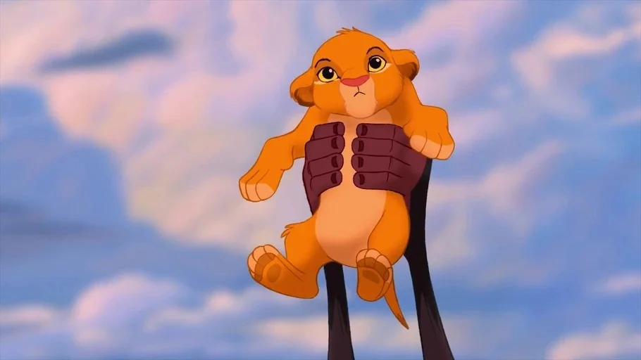 The Lion King (1994) movie still: closeup — A cartoon lion cub is held aloft against a soft, light blue cloudy sky. The cub is being lifted by two…