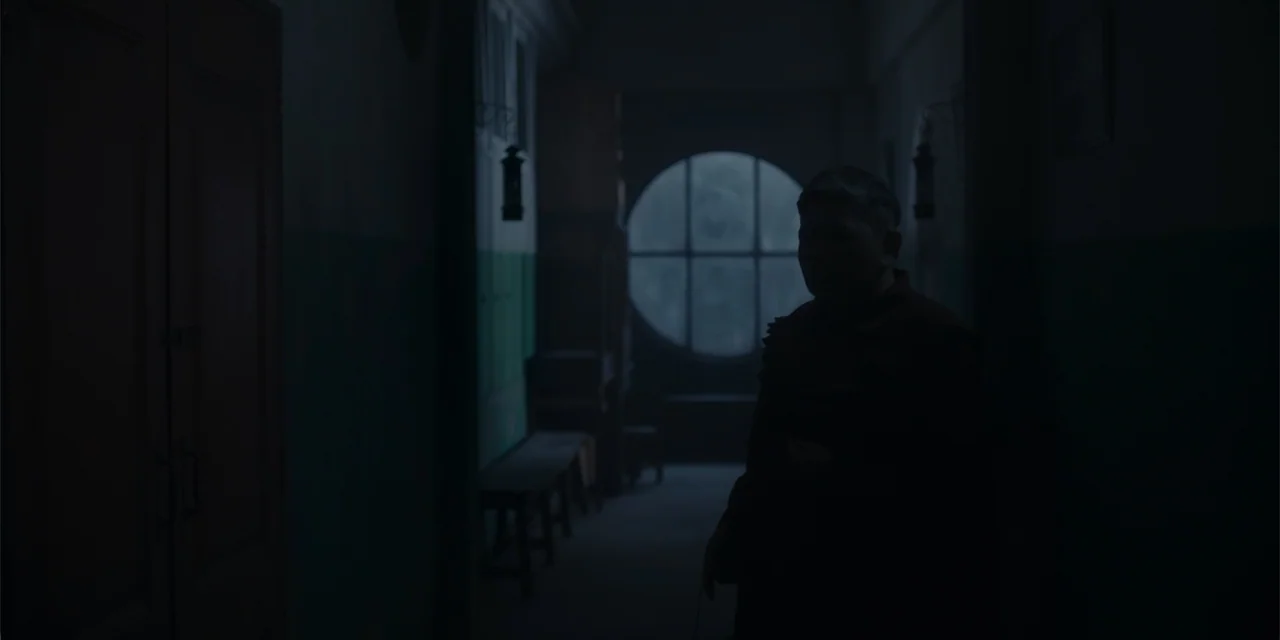 Host (2020) movie still: medium — A dark, dimly lit corridor features a large circular window at the end. A man stands in the foreground as a…