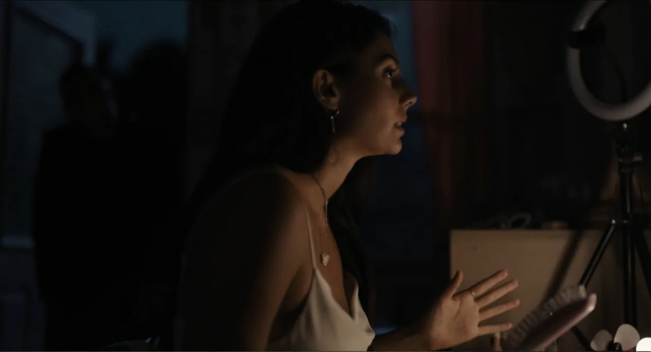 Hell of a Summer (2025) movie still: medium — A dark bedroom interior features a young woman lit by a bright ring light. The woman sits in the…