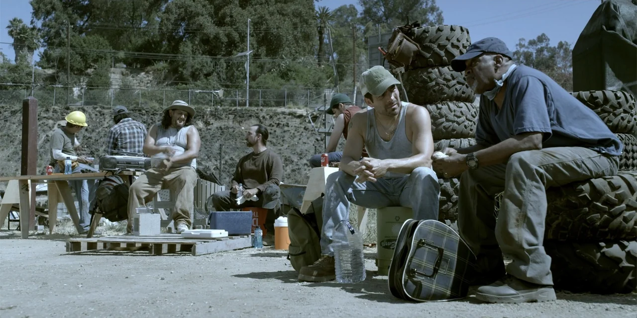 7th & Union (2021) movie still: medium — A dusty outdoor yard features stacks of tires and various tools near a fence. Two men sit together in the…