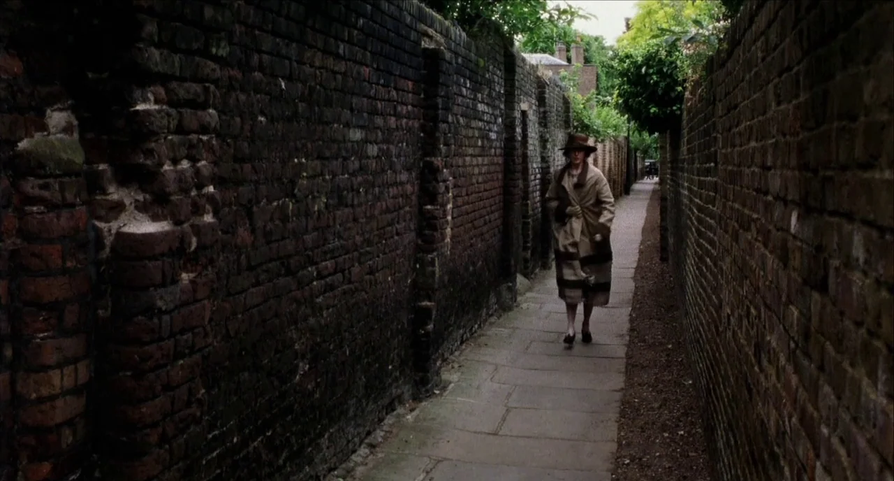 The Hours (2002) movie still: fullbody — A narrow stone-paved walkway is enclosed by tall, weathered brick walls on both sides. A woman in a brown…
