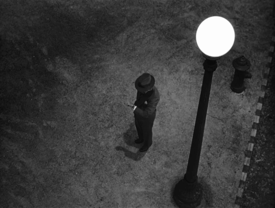 Strangers on a Train (1951) movie still: wide — A dark, paved area features a single lit streetlamp casting a circular glow. A man wearing a fedora…