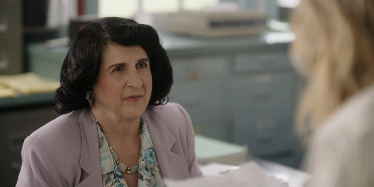 Totally Killer (2023) movie still: medium — A 1980s workplace with grey metal file cabinets and a cluttered desktop. A middle-aged woman sits at the…