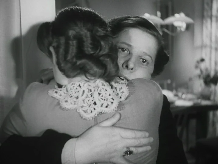 Crisis (1946) movie still: closeup — A domestic interior with out-of-focus furnishings visible in the background. Two women are embracing each other…