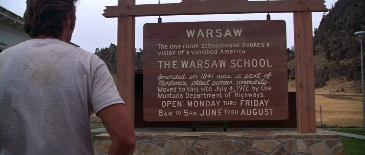 Thunderbolt & Lightfoot (1974) movie still: medium — A wooden informational sign stands in a dry, mountainous area with scrubby terrain. A man…