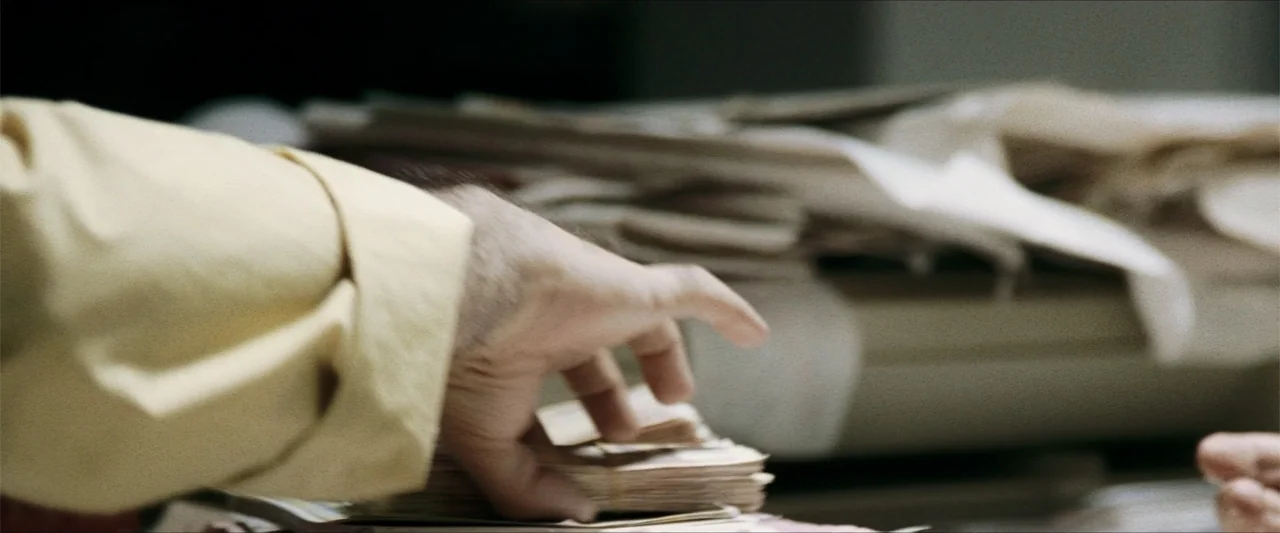 GOMORRAH (2023) movie still: closeup — A cluttered desk surface holds stacks of white paper and documents. A hand wearing a pale yellow shirt…