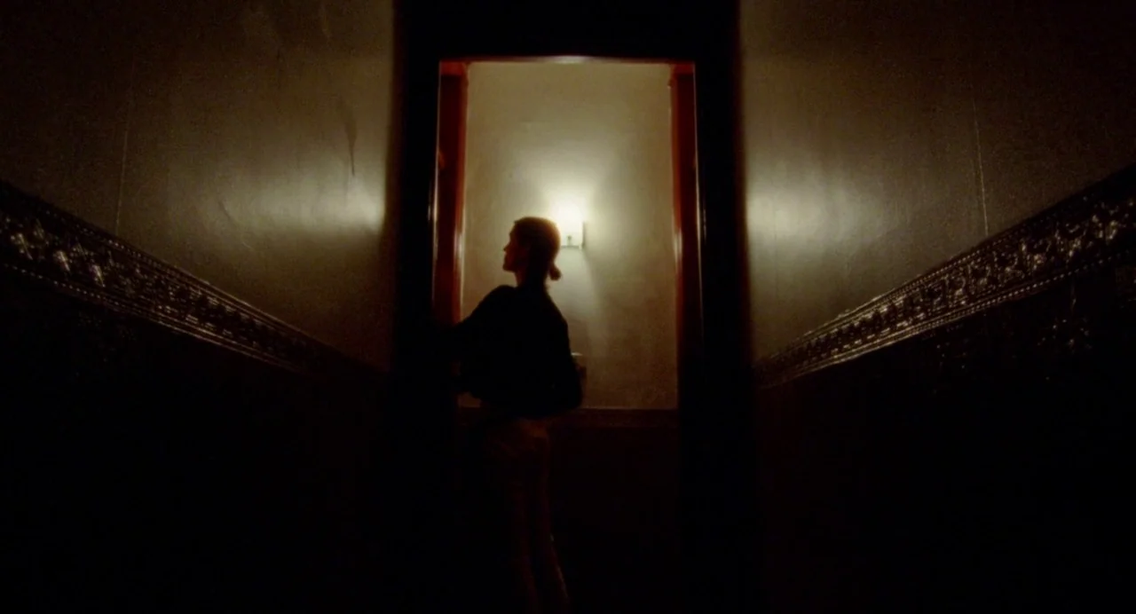 So Pretty (2019) movie still: wide — A dimly lit narrow hallway with decorative dark wainscoting and smooth upper walls. A woman stands as a…