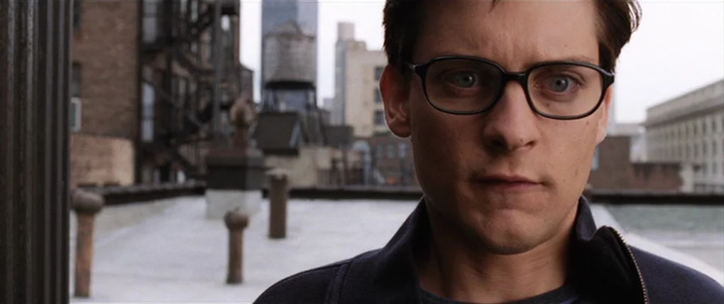Spider-Man 2 (2004) movie still: closeup — A flat urban rooftop with ventilation pipes and a distant water tower in view. A young man wearing…